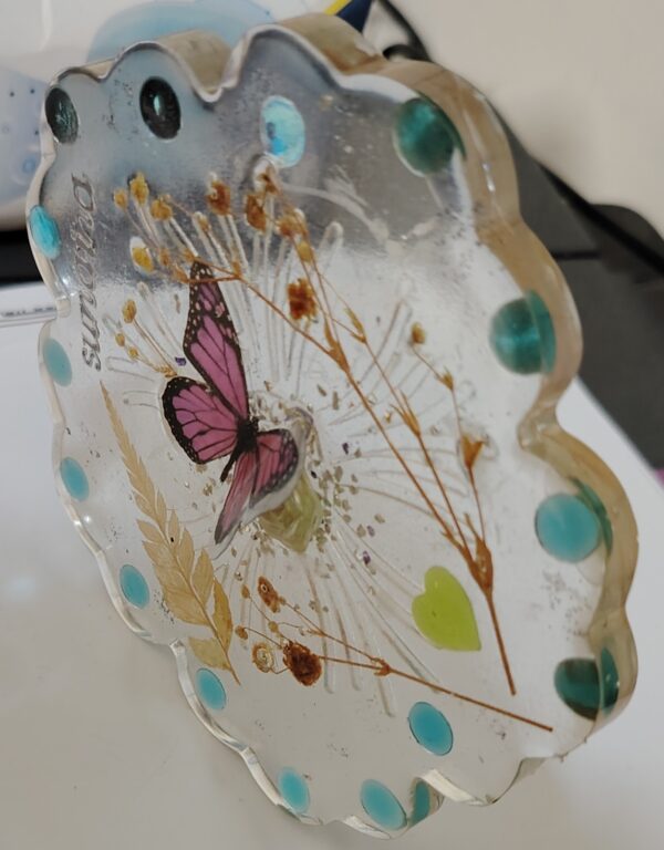 Handmade transparent resin coaster with pink butterfly, dried flowers, green heart, and turquoise dots